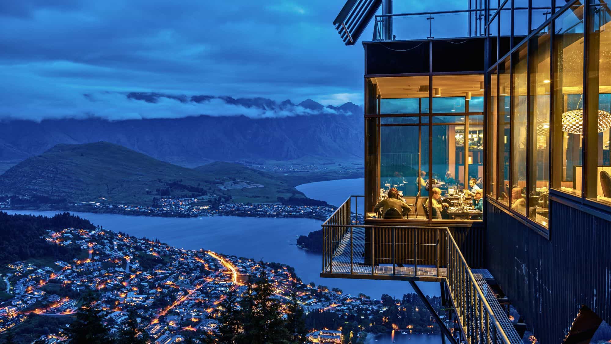 A modern glass-walled restaurant juts out from a mountainside, offering panoramic twilight views of Queenstown’s lights and the lake far below.