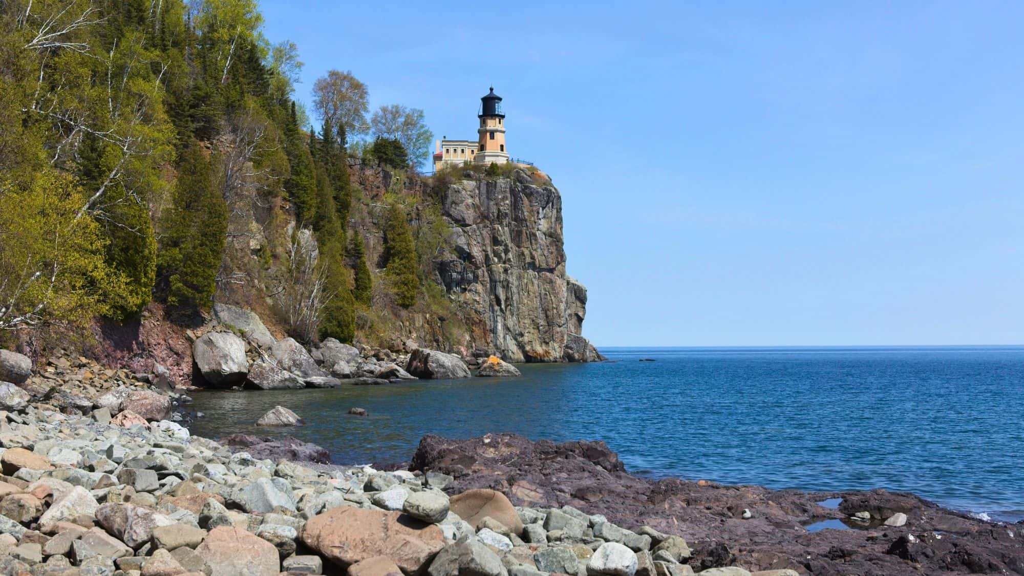 A lighthouse stands perched atop a sheer cliff on Minnesota’s North Shore, overlooking the deep blue expanse of Lake Superior with rocky shoreline below.