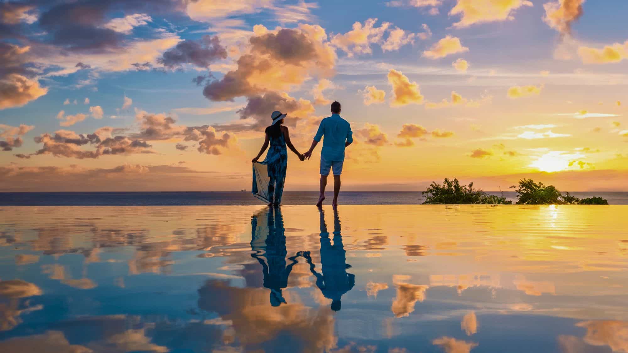 Framed by calm sea and scattered clouds, a couple stands barefoot at the edge of an infinity pool, their reflection blending with the glowing Caribbean sky.