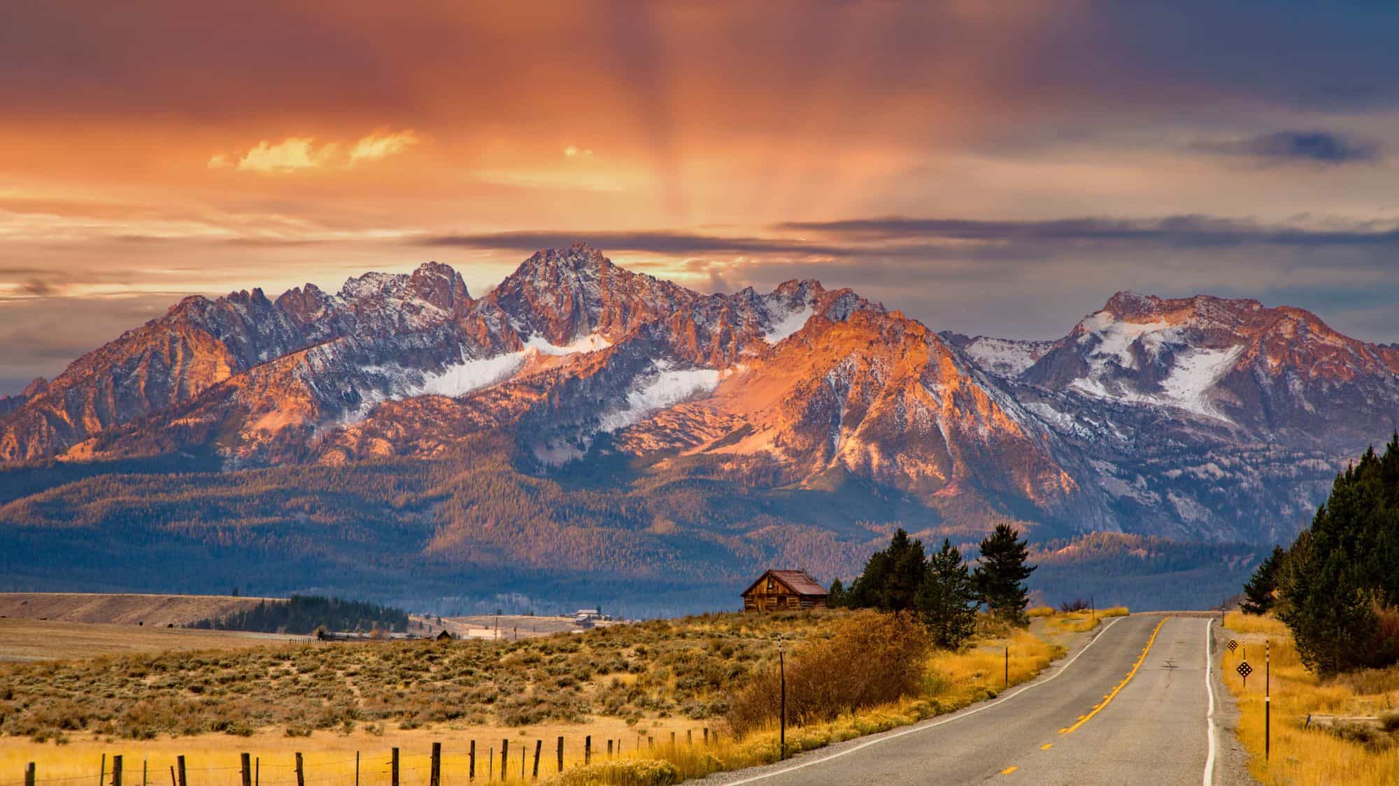 The Sawtooth Mountains glow under streaks of golden light as a quiet road leads toward rustic cabins nestled in the wide open valley near Stanley, Idaho.