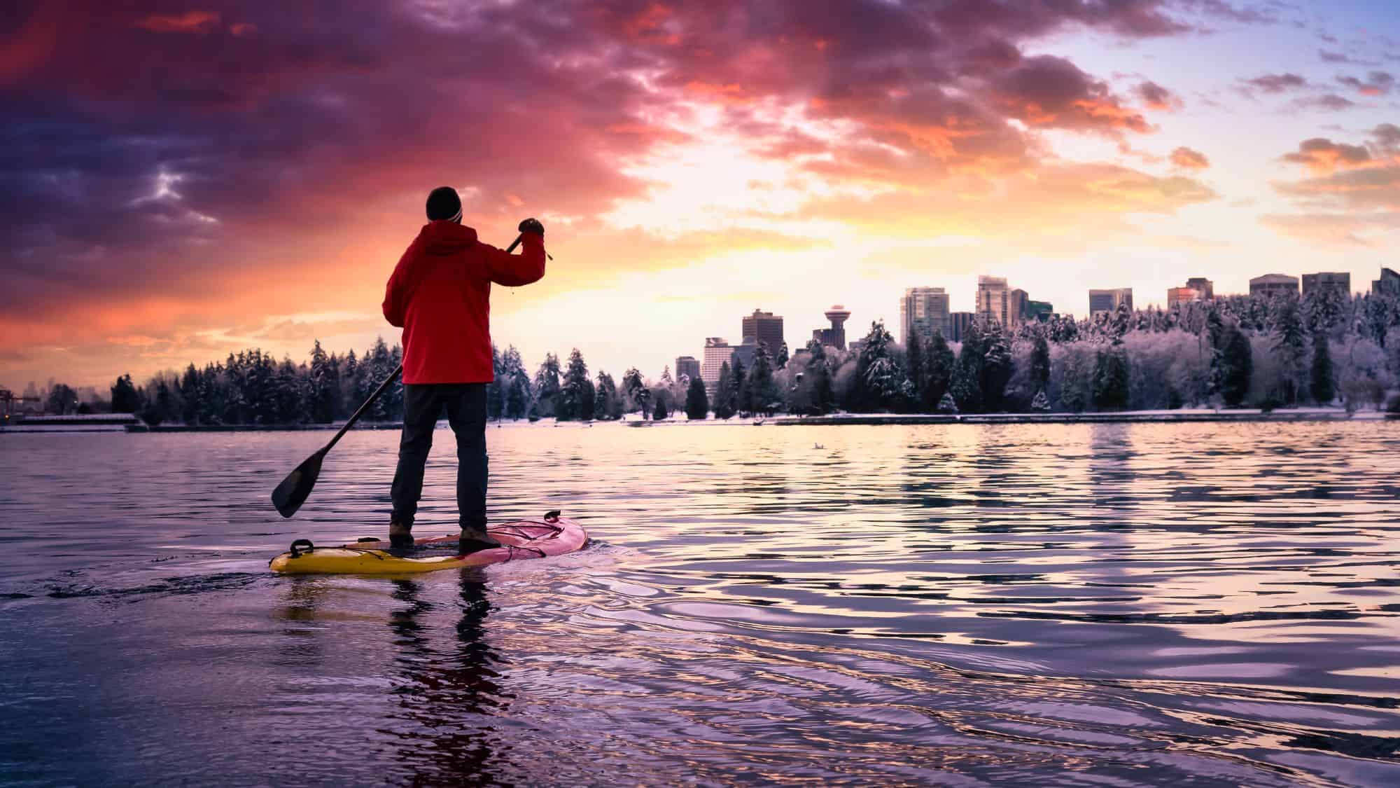 A paddleboarder glides across reflective waters as a snowy city skyline glows beneath a colorful winter sunset.