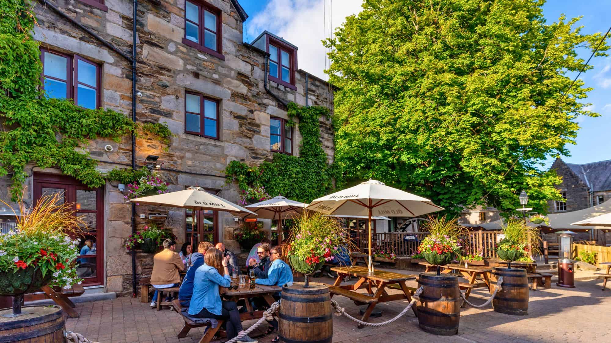 Stone walls draped in ivy surround a cozy beer garden with wooden tables, white umbrellas, and blooming flowers on a sunny afternoon.
