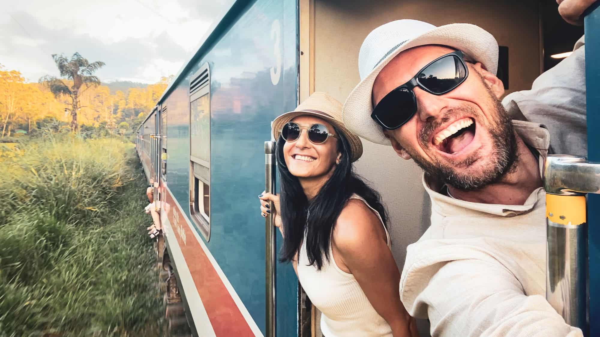 A couple beams with excitement as they lean out of a moving train, surrounded by lush greenery and golden light filtering through tropical trees.