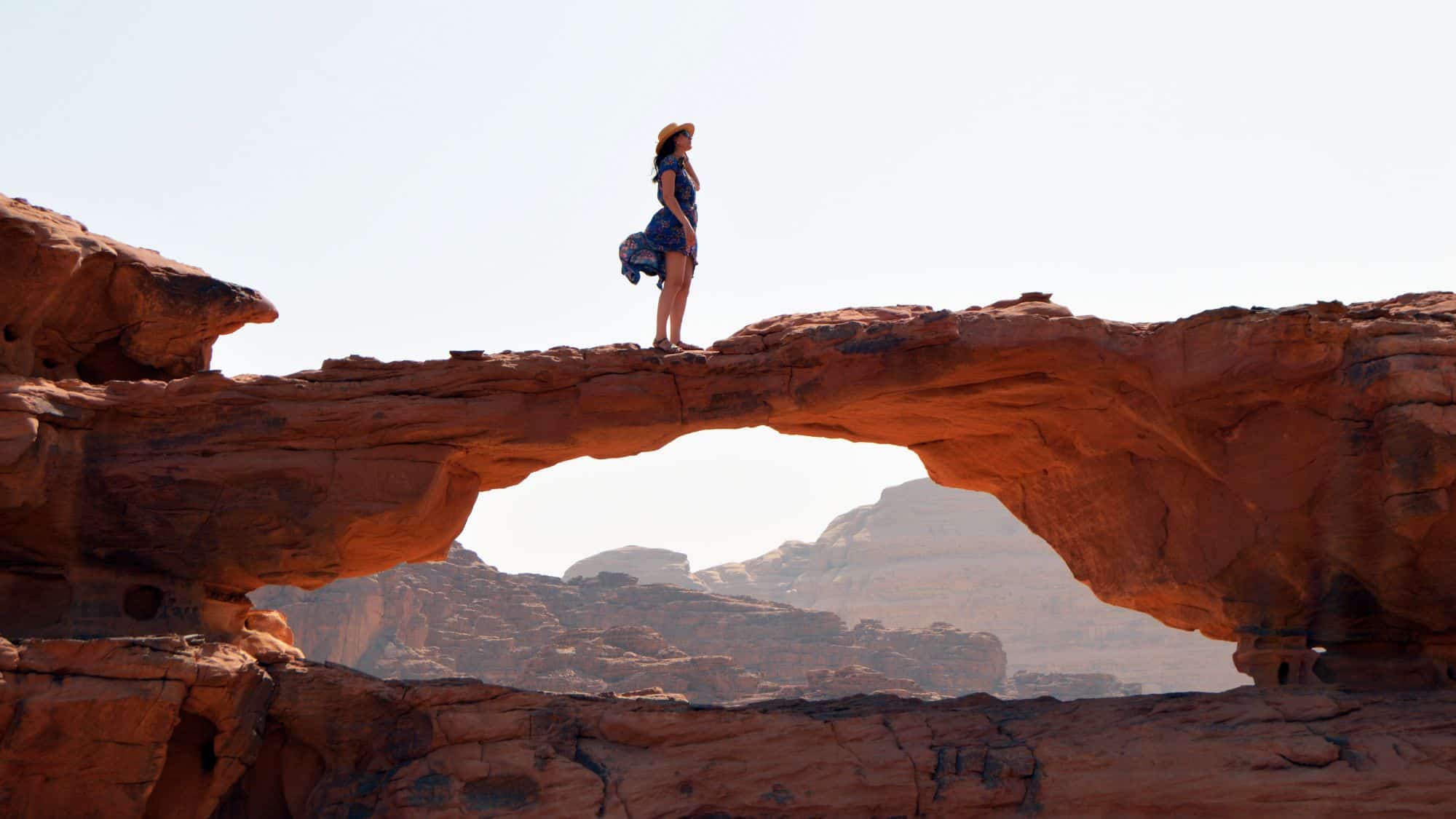 A woman in a summer dress stands atop a narrow sandstone arch, framed by layered red rock formations and a pale desert sky.