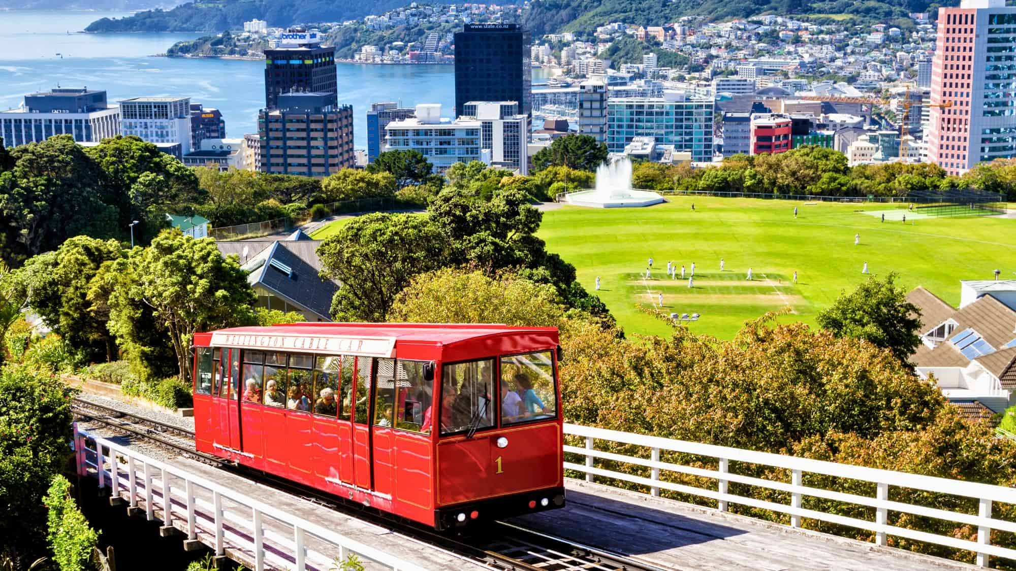 A bright red cable car ascends above the cityscape of Wellington, New Zealand, with lush gardens, urban buildings, and a glinting harbor below.