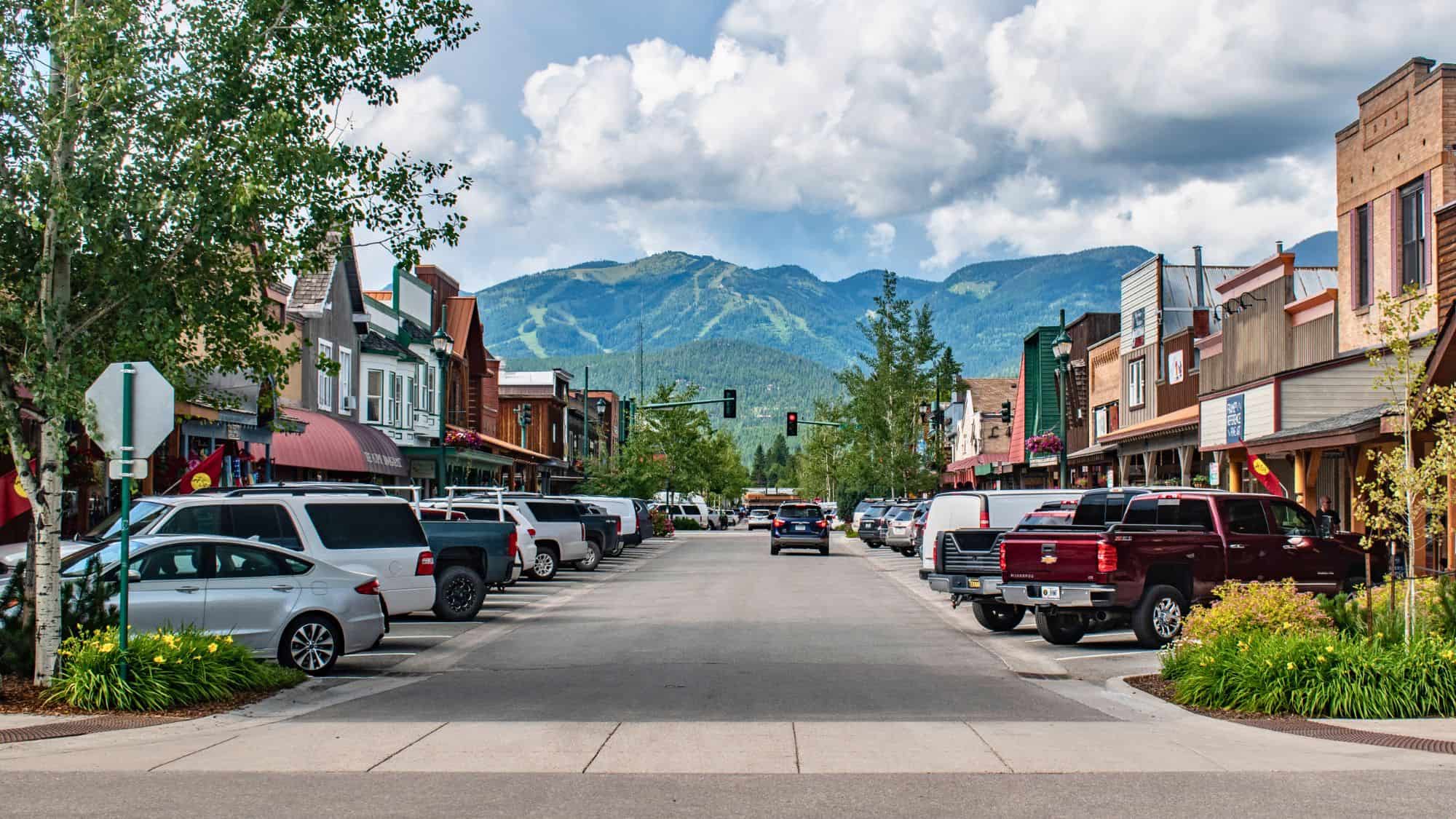A welcoming small-town street in Whitefish stretches toward forested ski slopes, with charming storefronts, parked trucks, and a backdrop of the Rocky Mountains.