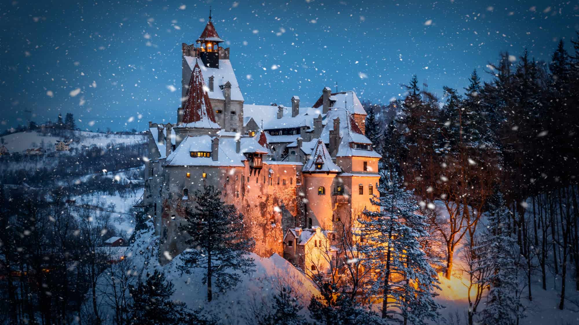 Snow falls over Romania’s Bran Castle, its warm lights glowing through the flurries as it rises dramatically from the forested hills.