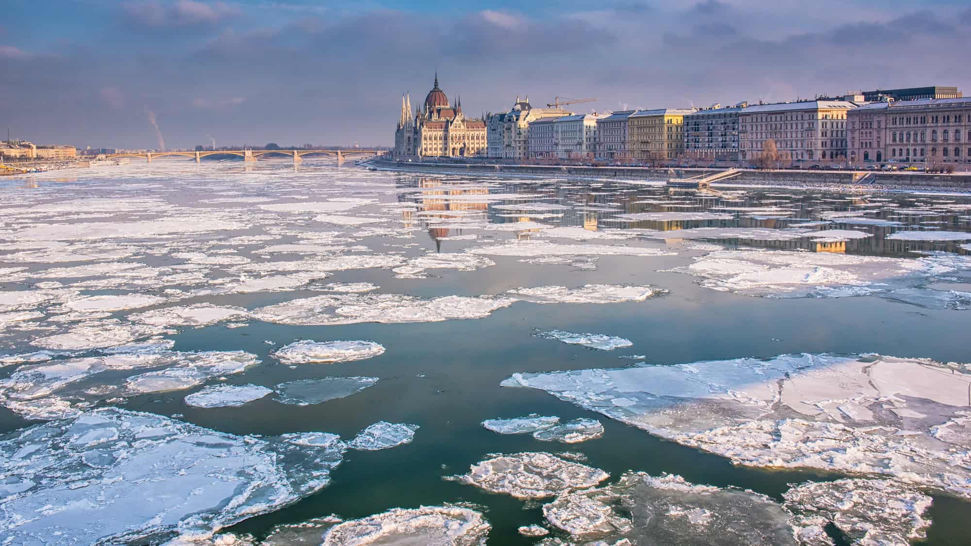 Large ice floes drift along the Danube River, reflecting the iconic Parliament building and surrounding cityscape under a pink-tinged winter sky.