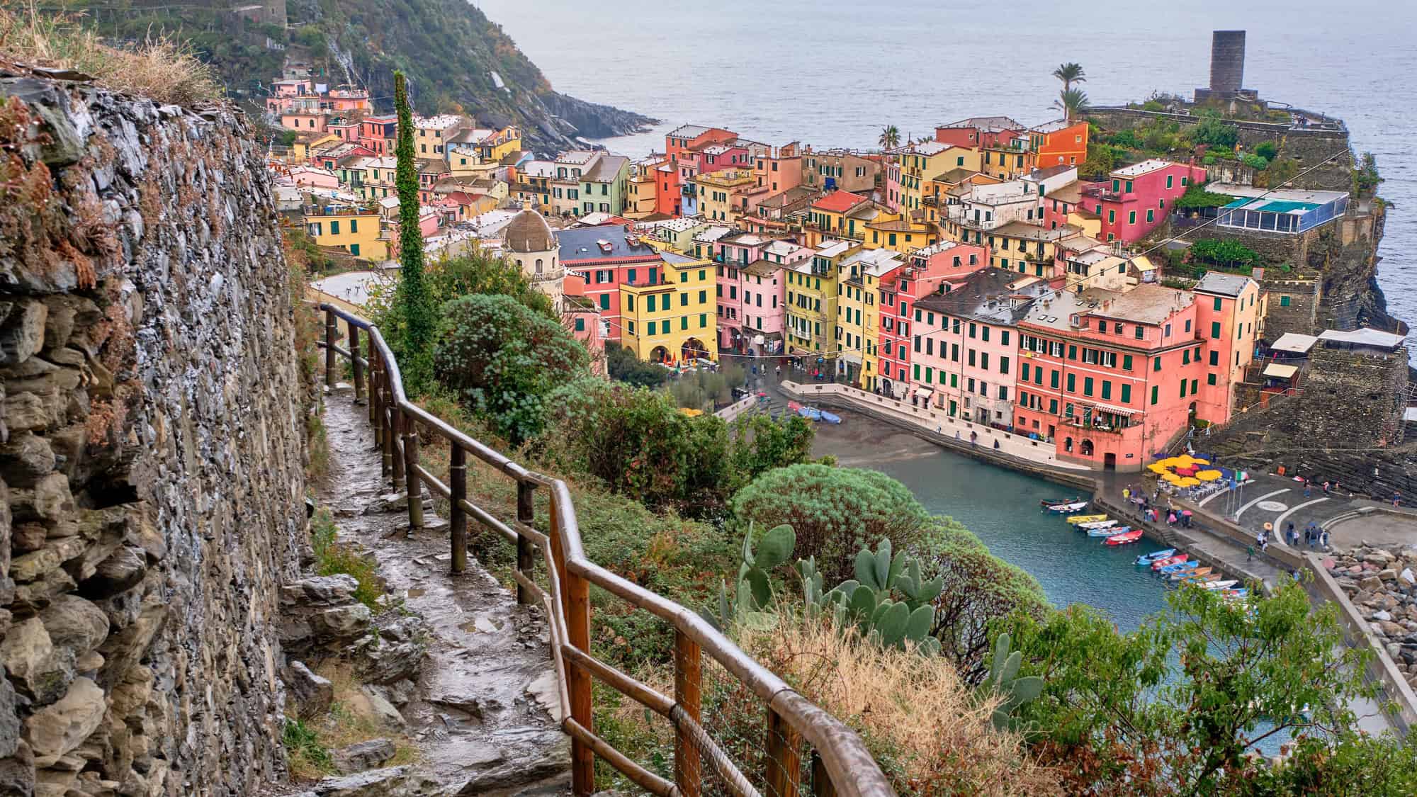 A narrow coastal trail leads to a colorful seaside village perched on the cliffs, its pastel buildings clustered tightly above a sheltered harbor.