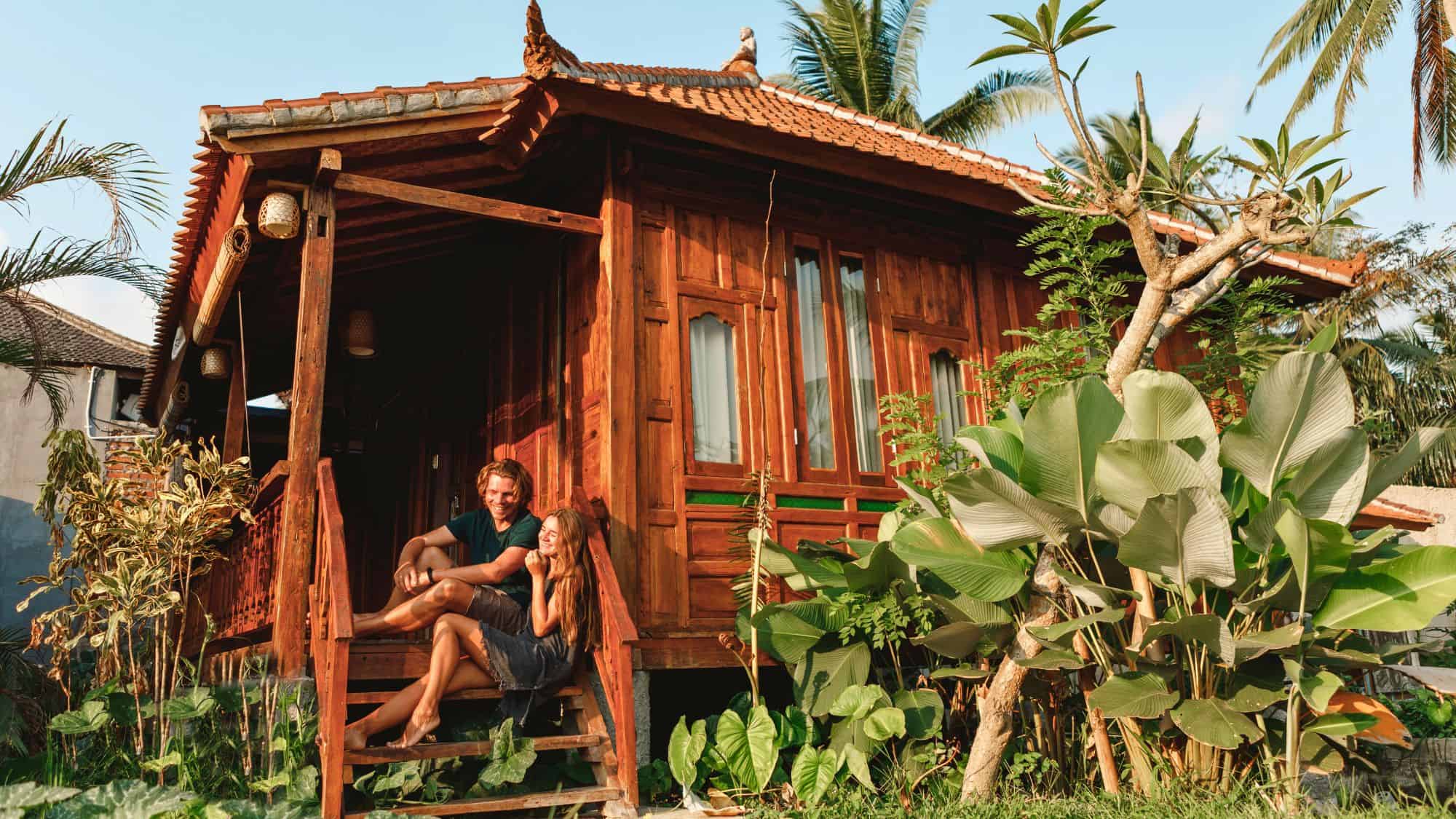 A couple relaxes on the steps of a traditional wooden house set among tropical plants and palm trees. The setting blends rustic charm with the laid-back feel of a tropical retreat.