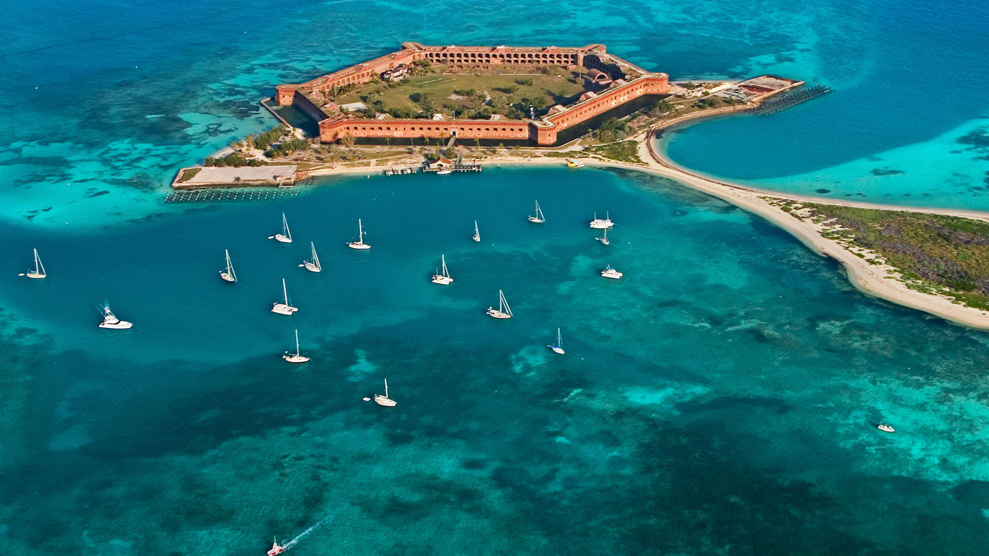 A cluster of sailboats surrounds Fort Jefferson, a massive coastal fortress set on a remote island with strikingly clear blue waters and coral shallows.