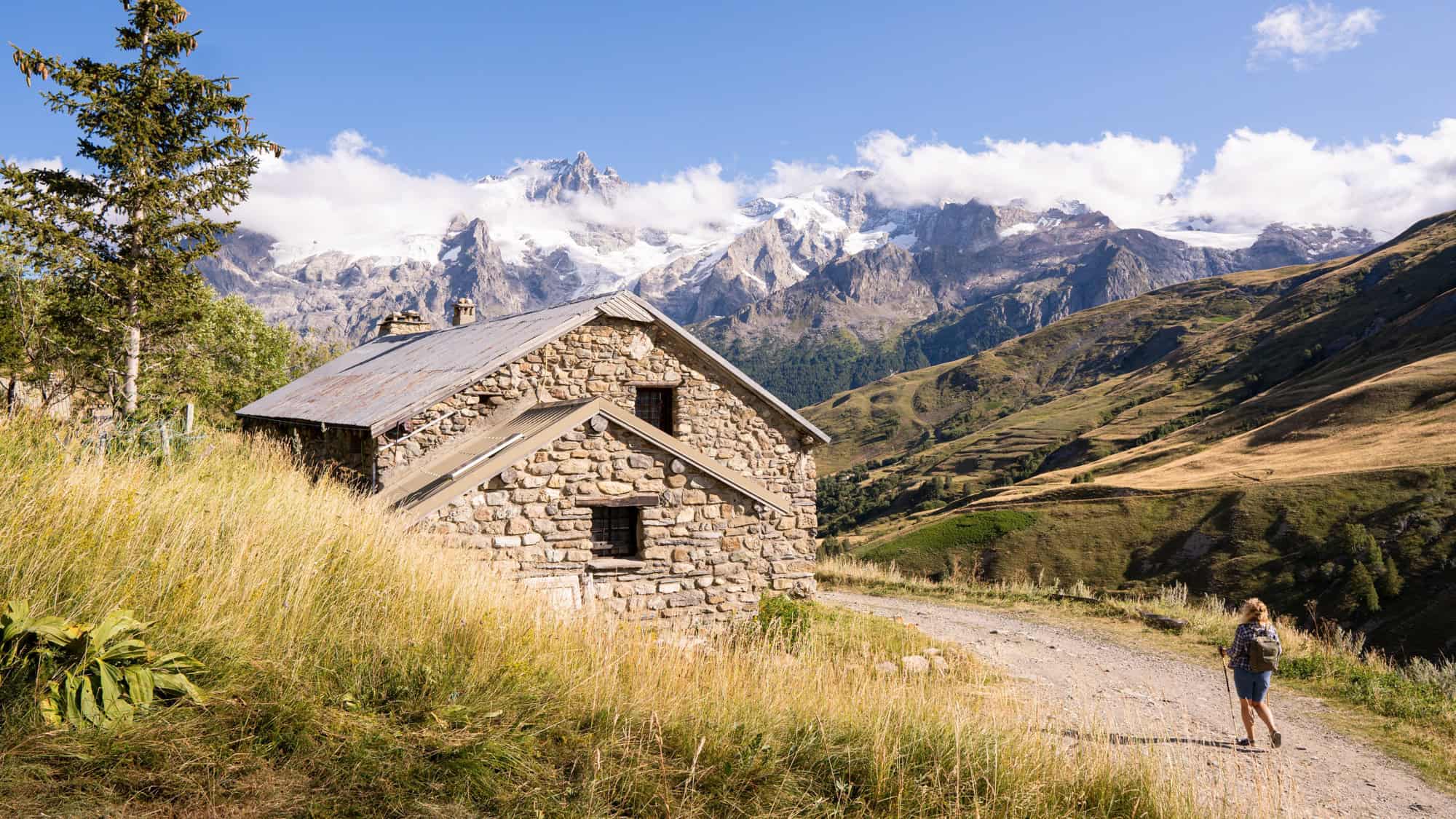 A hiker walks past a rustic stone hut set in tall golden grass, with sweeping views of the French Alps rising sharply in the distance.