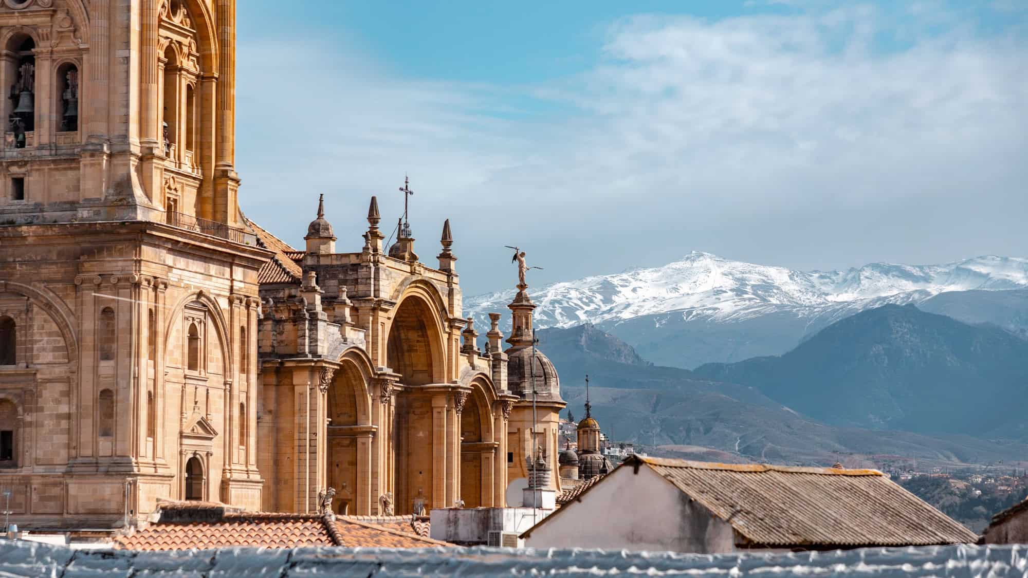 The intricate sandstone facade of Granada Cathedral stands against a backdrop of snow-dusted peaks in southern Spain’s Sierra Nevada range.