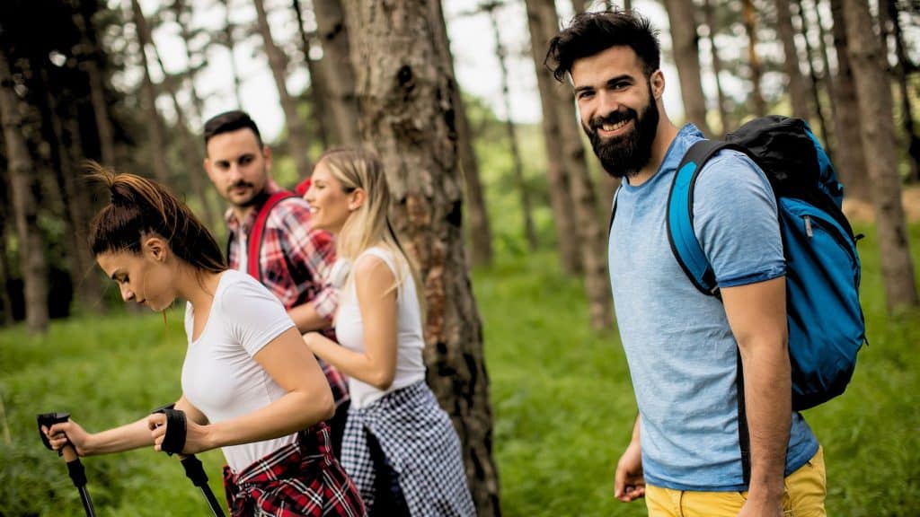 Four hikers move through a sun-dappled pine forest, one smiling at the camera while the others concentrate on the trail ahead.