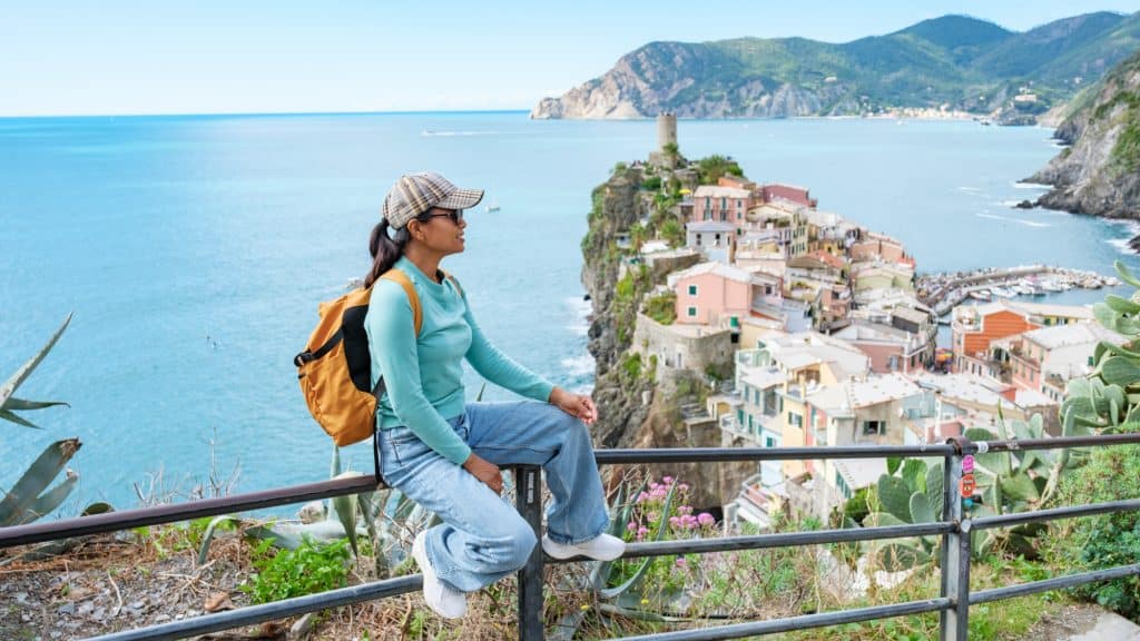 A traveler rests along a coastal viewpoint gazing over colorful hillside homes stacked above the Ligurian Sea, framed by mountains and turquoise water.