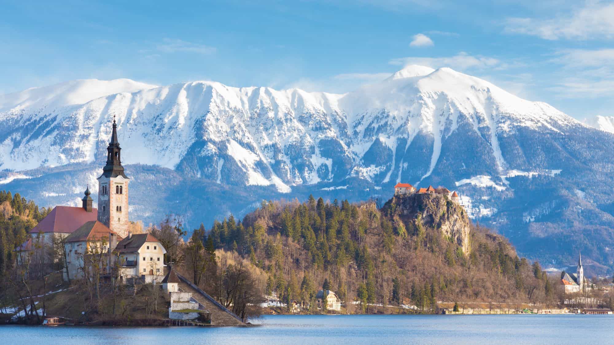 A picturesque church sits on an island in Lake Bled, backed by dramatic snow-covered mountains and a hilltop castle in the distance.