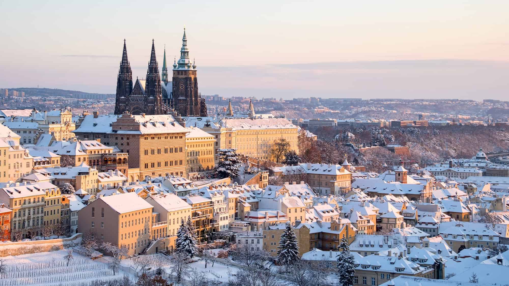 The rooftops of Prague are blanketed in snow, with the spires of St. Vitus Cathedral rising above the Old Town at sunrise.