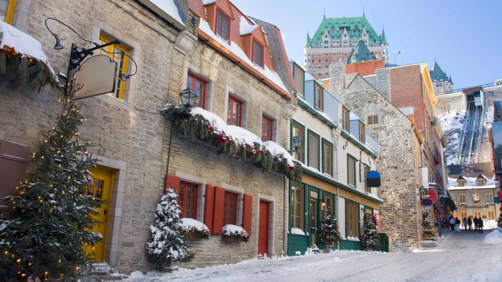 Colorful stone buildings line a narrow, snowy street in Old Quebec, with holiday decorations and Château Frontenac towering in the background.