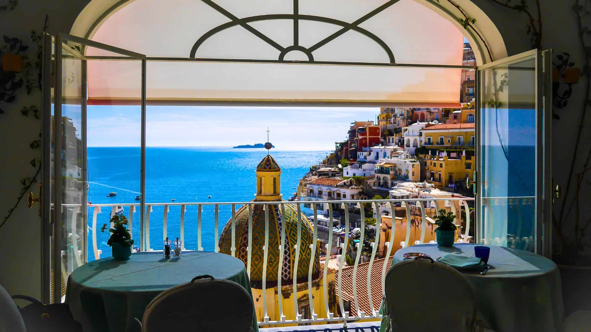 Open French doors frame a romantic terrace overlooking Positano’s cliffside homes and the bright blue sea, with a tiled church dome in the foreground.