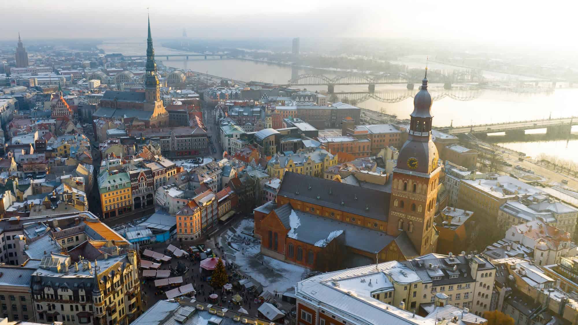The towers of Riga rise over pastel-colored rooftops dusted with snow, as the city stretches toward the Daugava River and misty horizon.