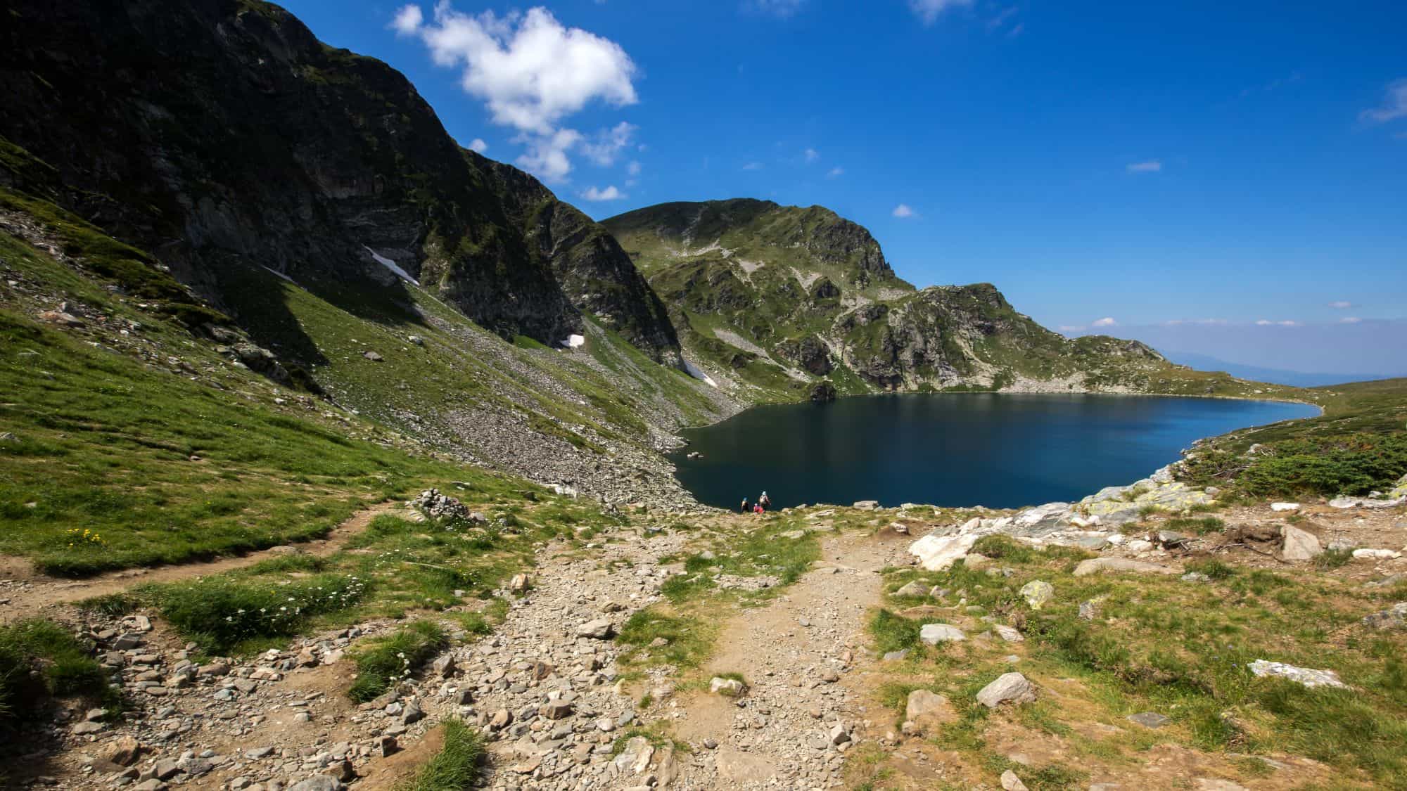 A rocky mountain trail leads down to a deep blue alpine lake nestled beneath green slopes and jagged cliffs under a wide open sky.