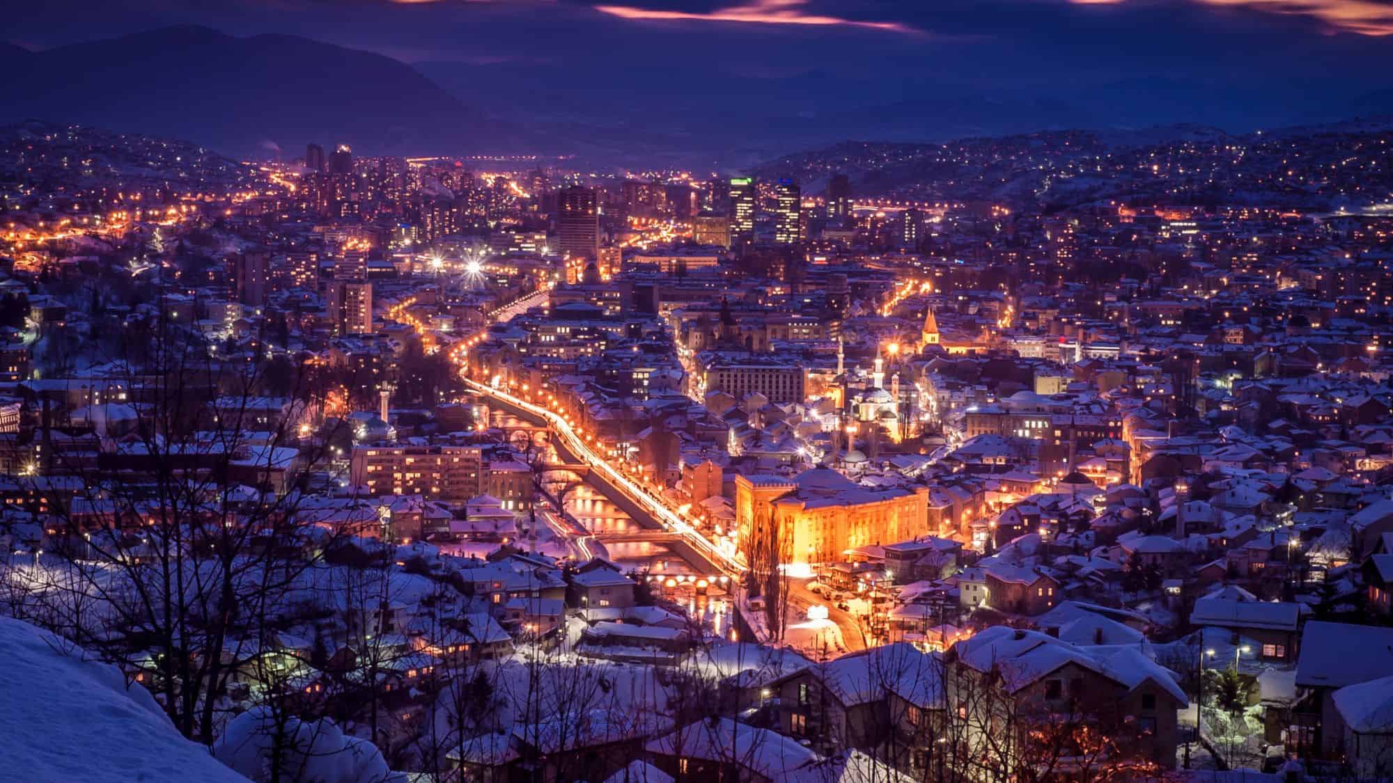 A golden ribbon of lights winds through snowy Sarajevo, as warm city lights glow against the deep purples and blues of twilight.