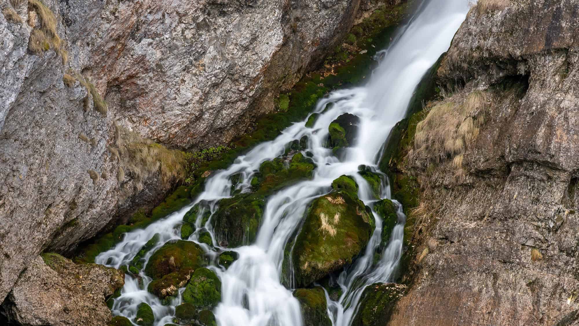 A thin cascade flows gently over moss-covered boulders and rugged stone, creating a tranquil natural scene tucked into a mountain slope.