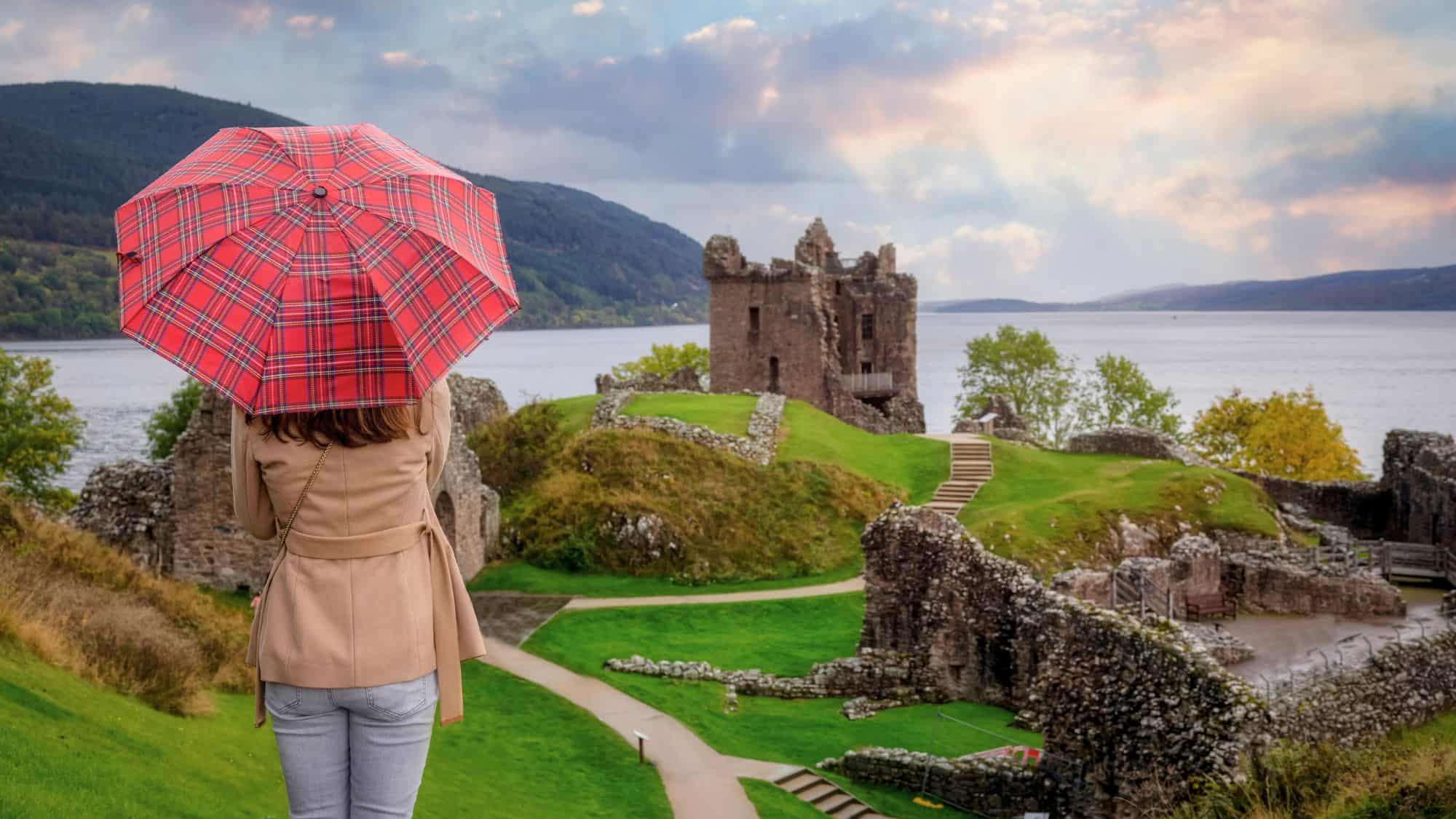 A person with a tartan umbrella looks out over the ruins of a medieval castle on the banks of a misty lake, framed by green hills and soft light.