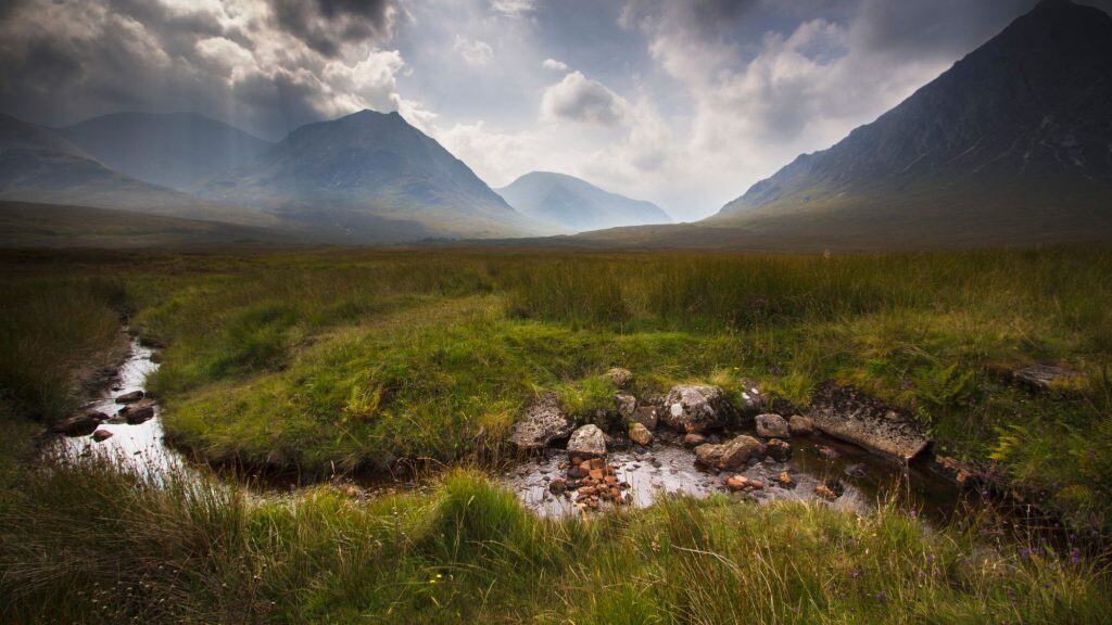 A dramatic dark day in the scottish highlands with a small stream flowing in front of tall mountains.