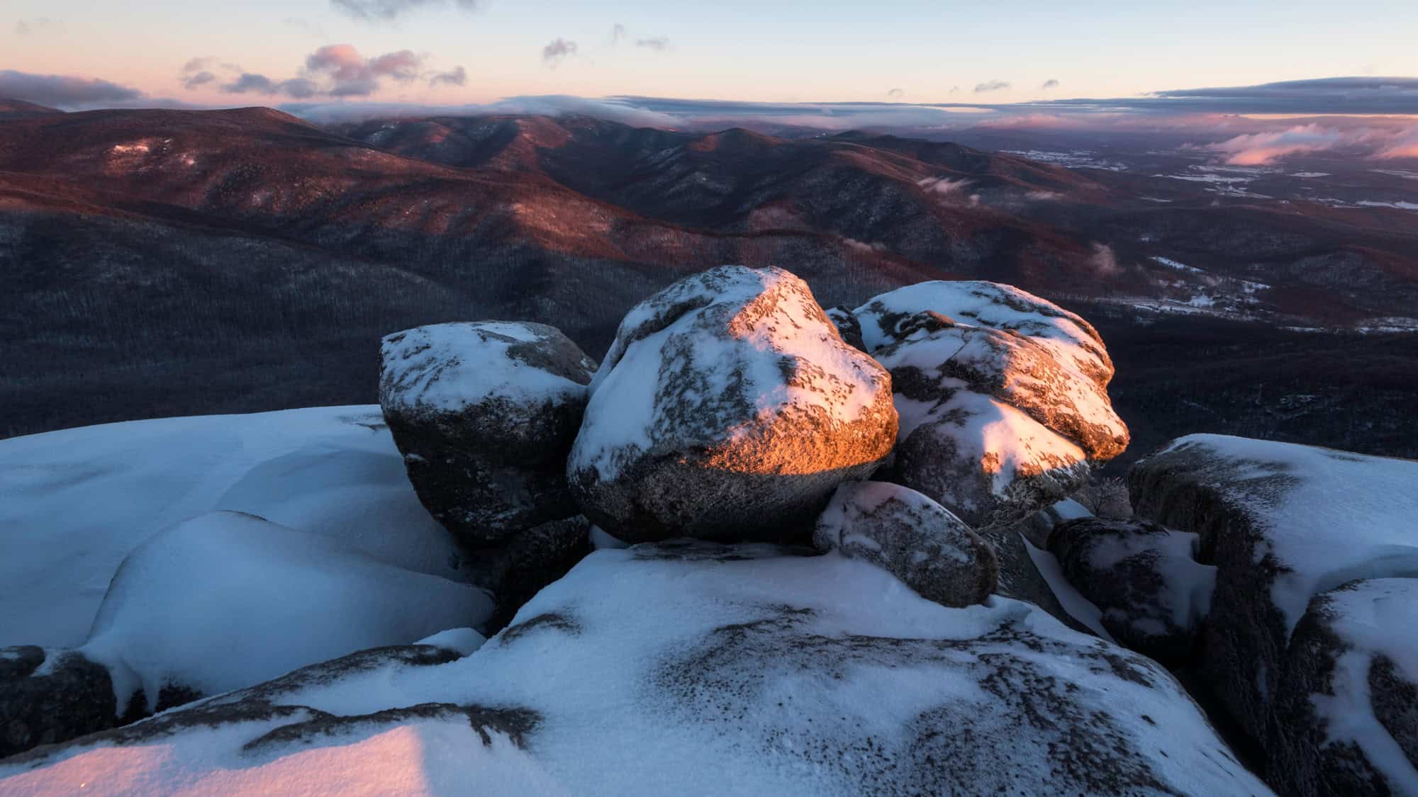 Large boulders topped with snow catch warm light at sunset, overlooking a stretch of softly shadowed ridges.