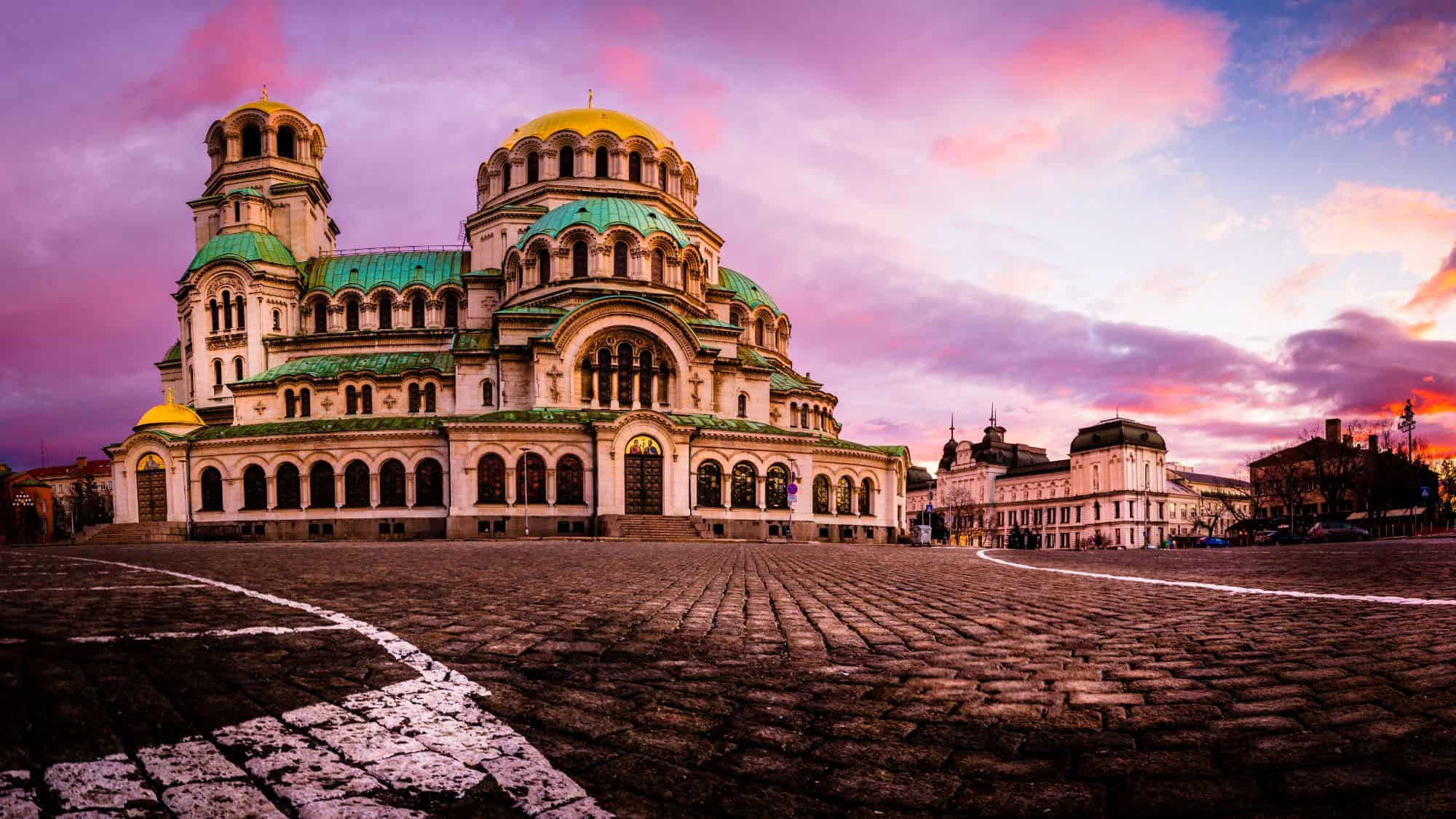 The Alexander Nevsky Cathedral in Sofia stands majestic under a vivid pink and purple sky, its domes glowing in the fading light.