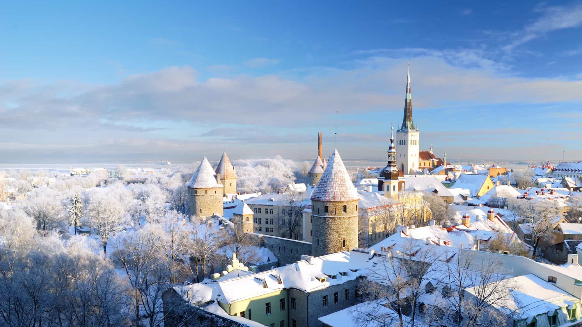 Medieval stone towers and pointed spires rise from a snowy old town under a bright blue sky, creating a fairytale scene in Estonia’s capital.
