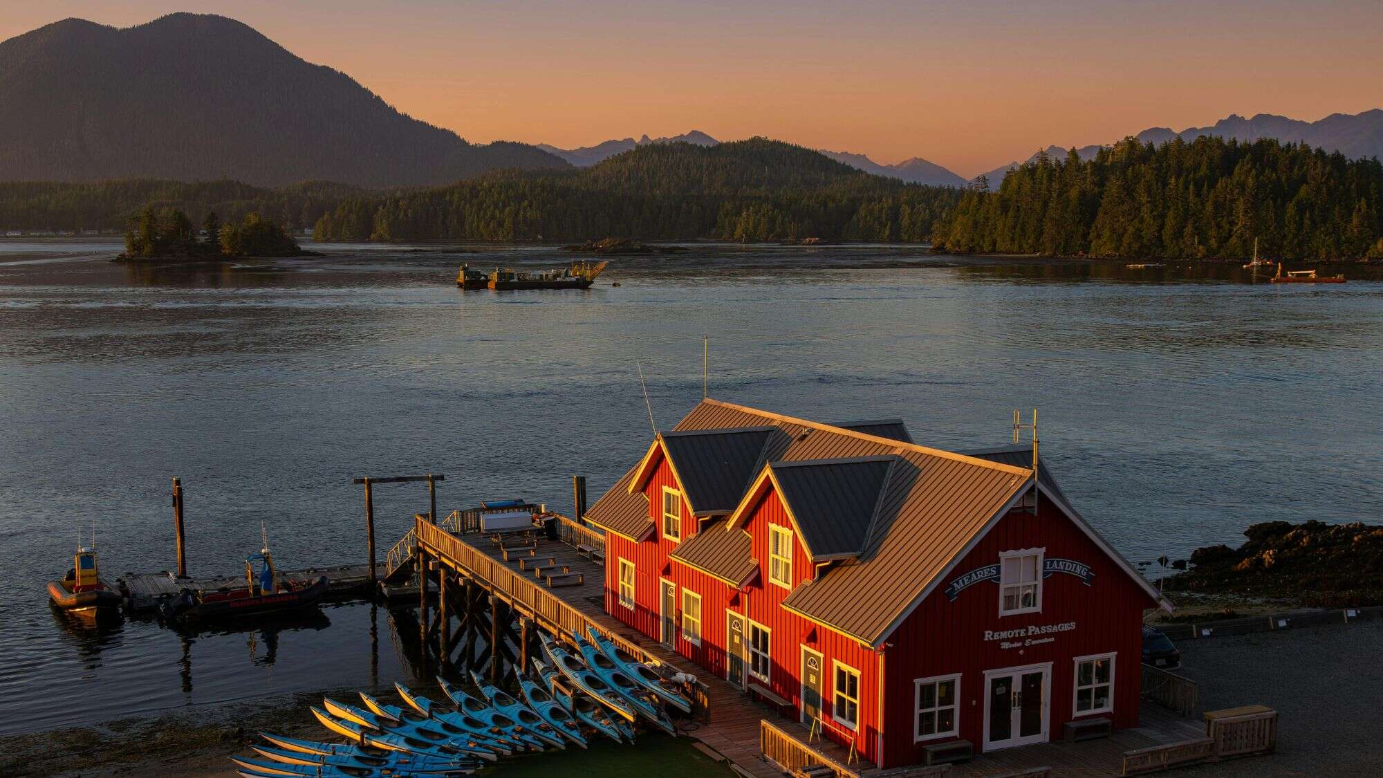 A bright red waterfront building sits at the edge of calm waters, with rows of blue kayaks ready for use and forested islands silhouetted against a golden evening sky.