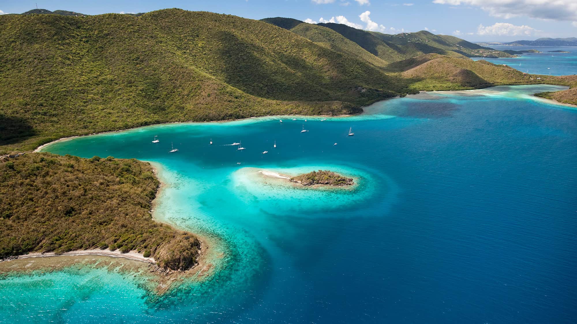 Turquoise waters and lush green hills frame this serene bay, dotted with sailboats and a tiny sandy islet—ideal for snorkeling or simply taking in the tropical views.