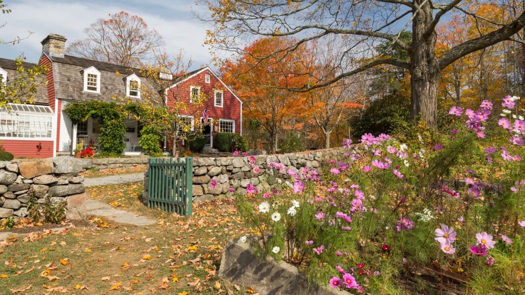A vibrant autumn setting featuring a red house with white trim, surrounded by colorful fall foliage, a stone wall, and a garden of pink and purple cosmos flowers. A green gate and dirt path lead to the house, adding to the charm.
