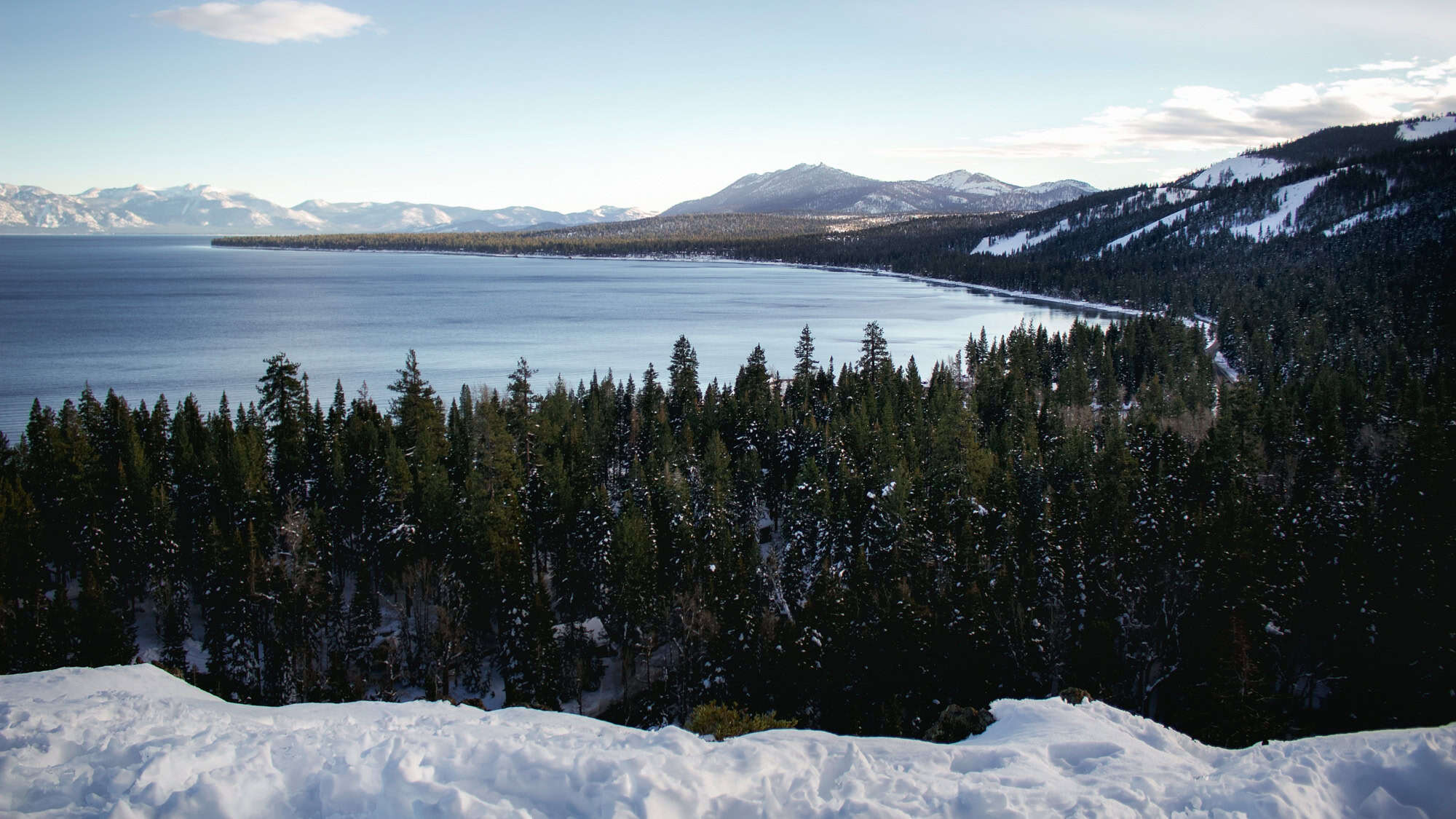 A sweeping view of snow-dusted pine forests and the deep blue waters of Lake Tahoe, framed by distant mountain ridges.