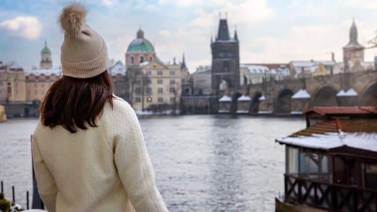 A woman in a cozy winter hat stands at the Vltava River’s edge, gazing at the historic Charles Bridge and Prague’s snow-dusted skyline.
