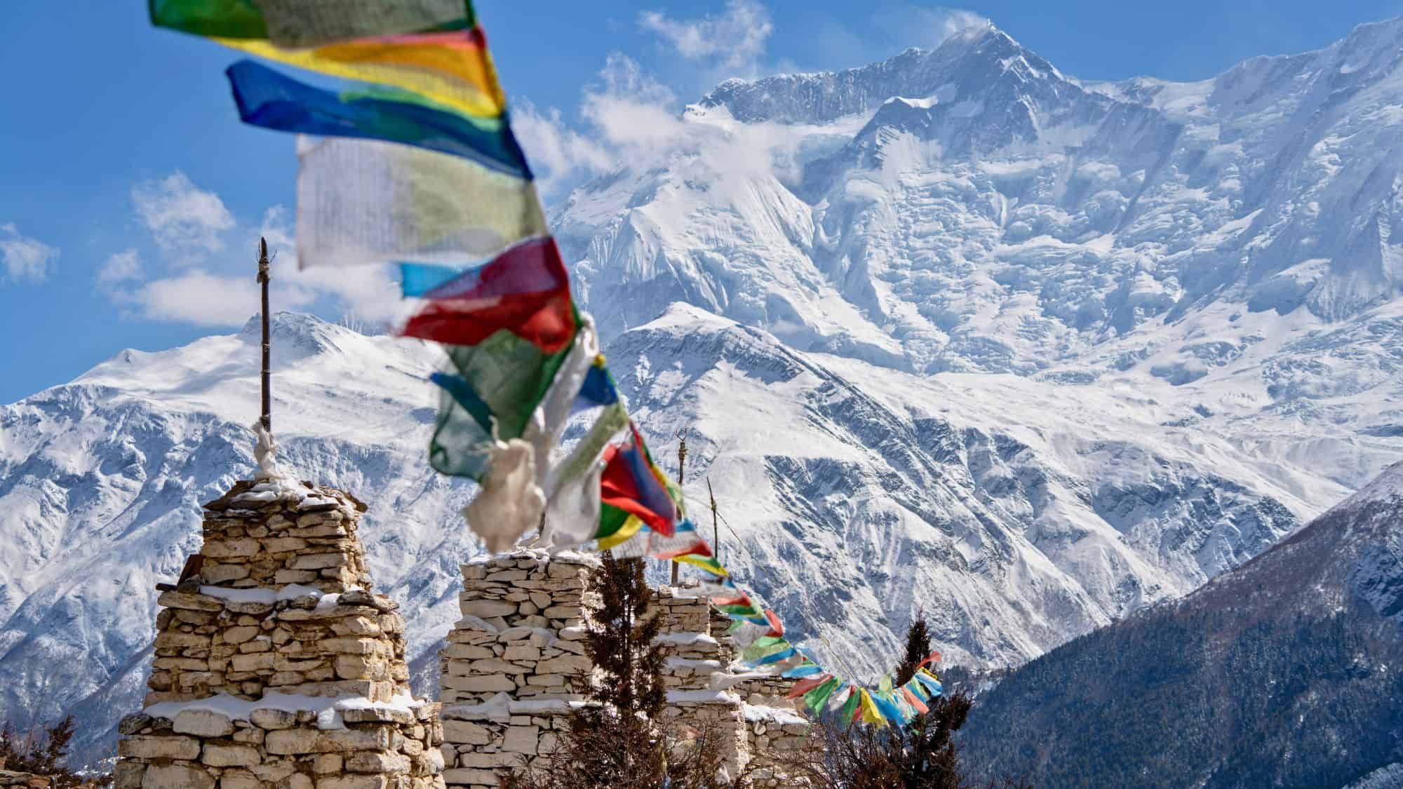 Colorful prayer flags flutter beside stone stupas in the foreground, set against towering, snow-covered Himalayan peaks under a clear blue sky.
