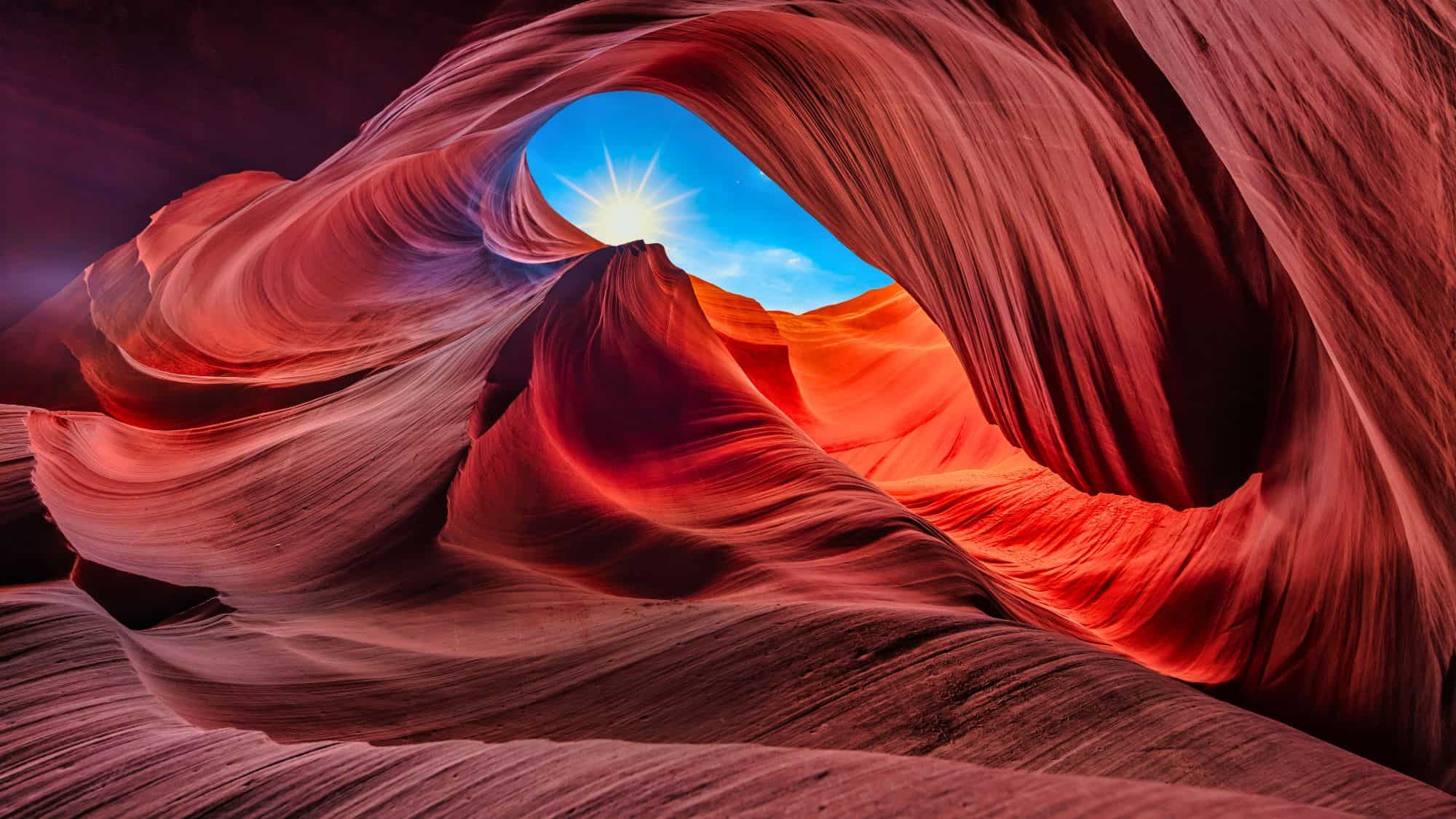Softly sculpted sandstone walls swirl upward in vibrant reds and oranges, framing a sunburst shining through a natural archway against a bright blue sky.