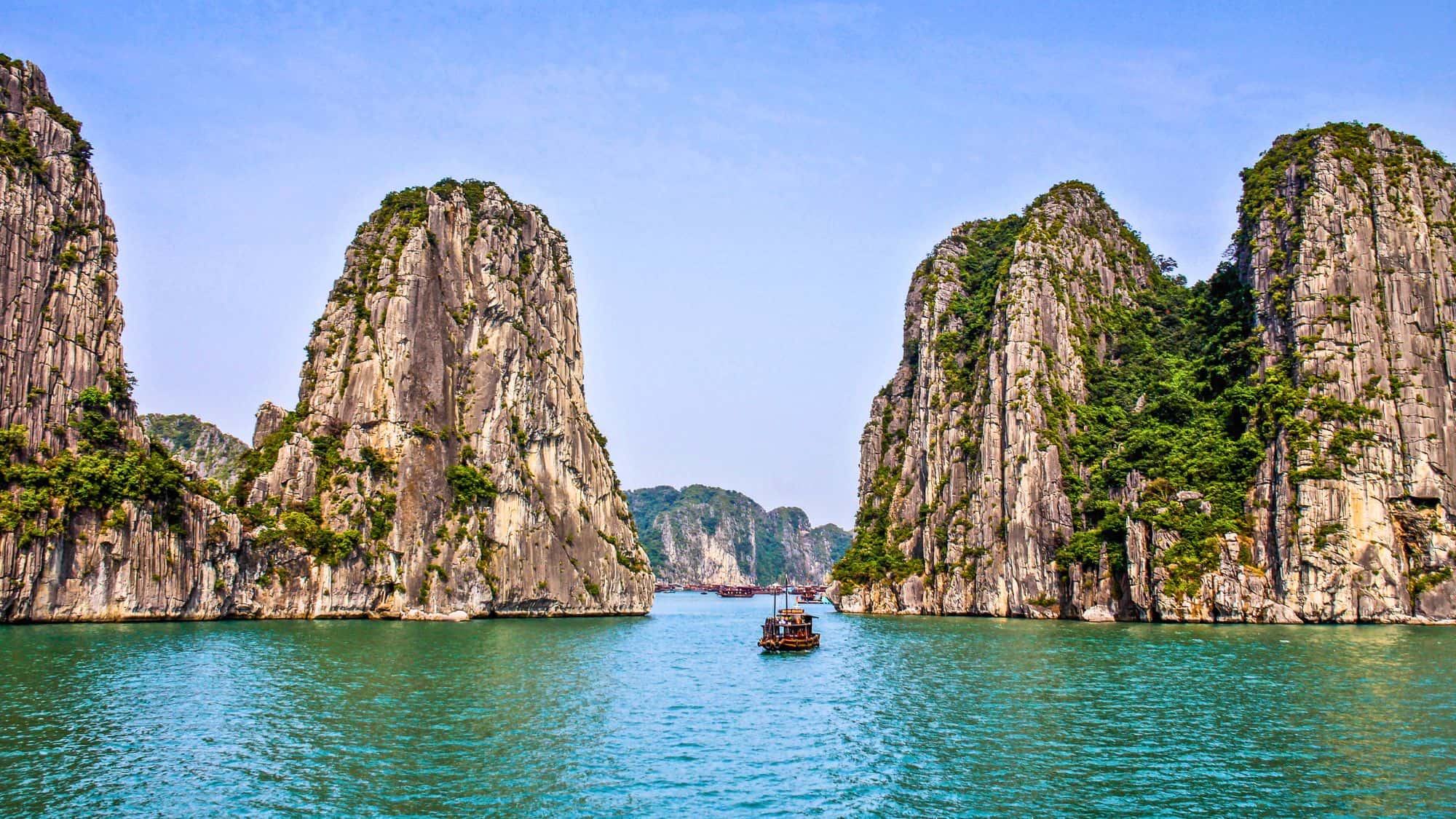 A traditional wooden boat sails between steep limestone karsts rising dramatically from emerald green water, with dozens more peaks visible in the distance.