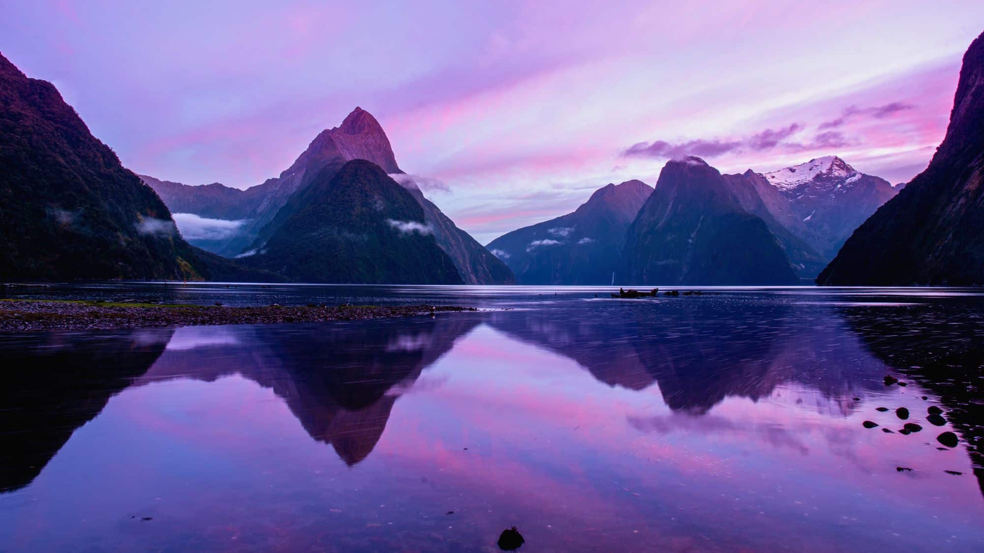 Jagged peaks of New Zealand’s Fiordland reflect in still waters under a violet and pink sky as twilight sets in.