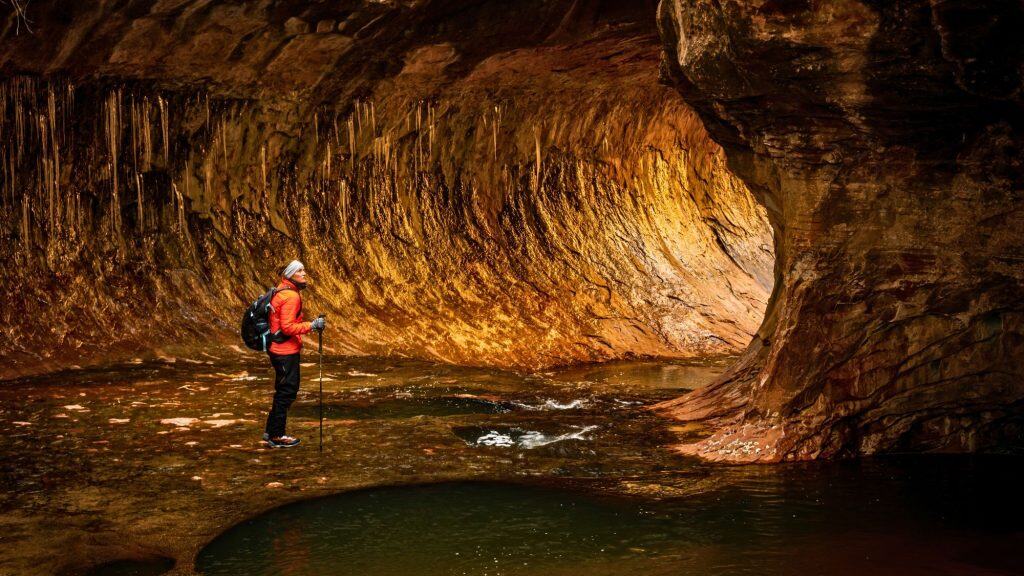 A solo hiker in a red jacket stands ankle-deep in clear water beneath a curving sandstone overhang, where warm rock tones glow in low canyon light and thin icicles hang from the ceiling. The scene emphasizes scale and stillness inside a narrow canyon shaped by flowing water.
