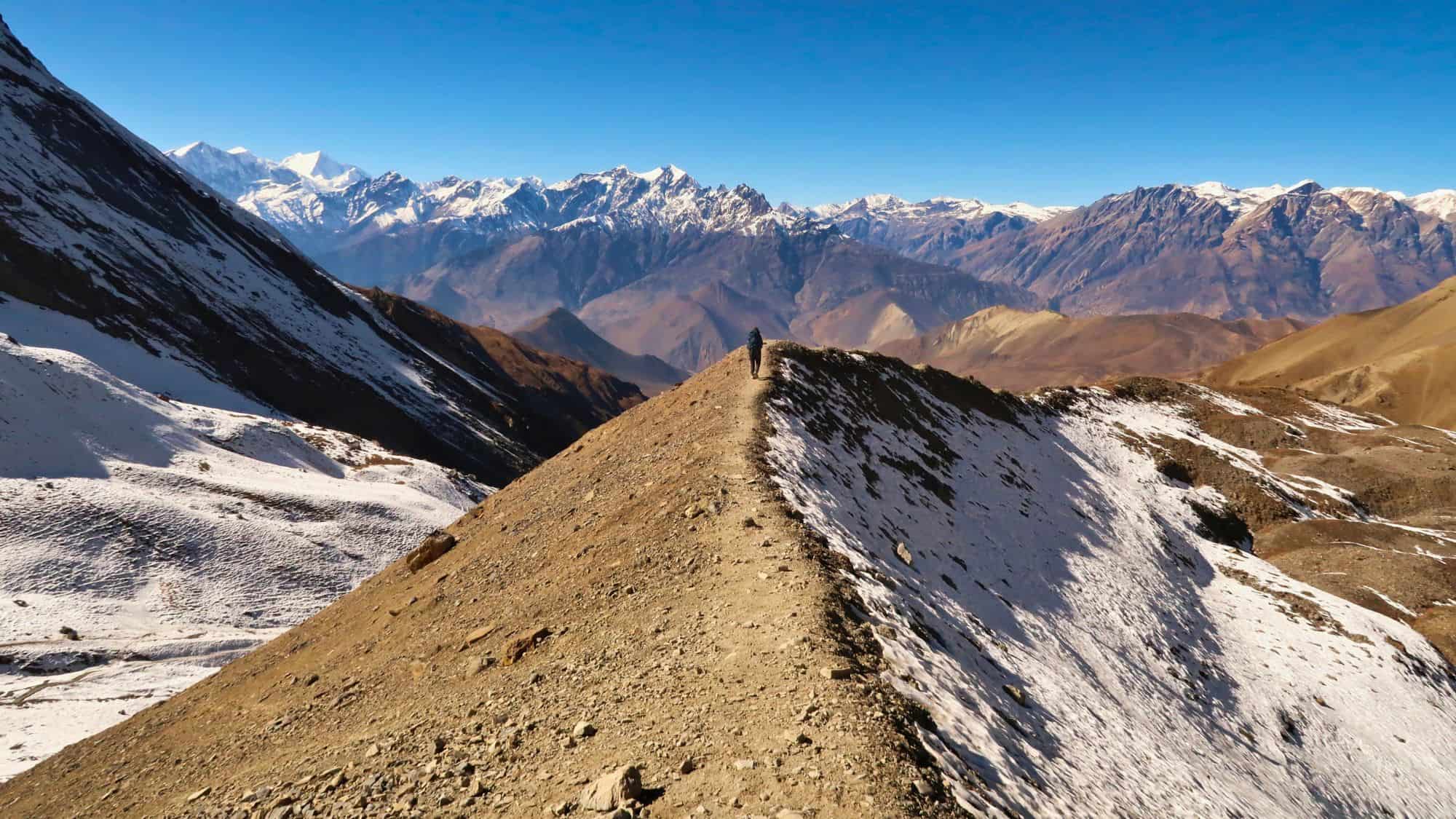 A lone trekker walks along a narrow ridge high in the Himalayas, surrounded by snow-dusted slopes and dramatic, distant peaks.