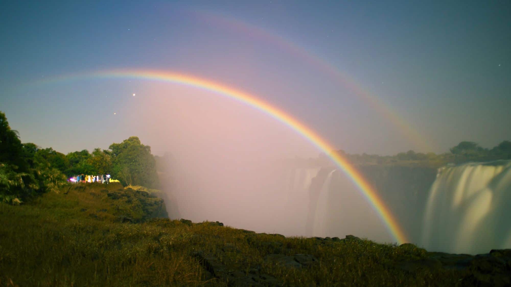 A vibrant moonbow arcs across the misty air above Victoria Falls, with its bands of color glowing softly against the nighttime sky.