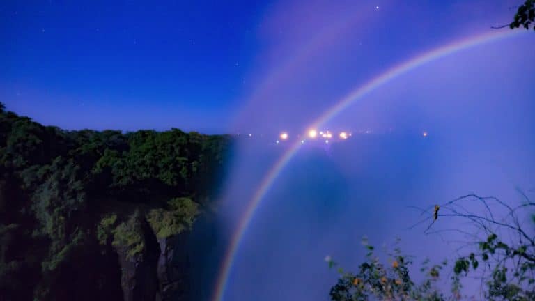 A moonbow arcs over a misty waterfall at night, framed by a starry sky, dense forest, and distant lights, creating a serene and magical scene.