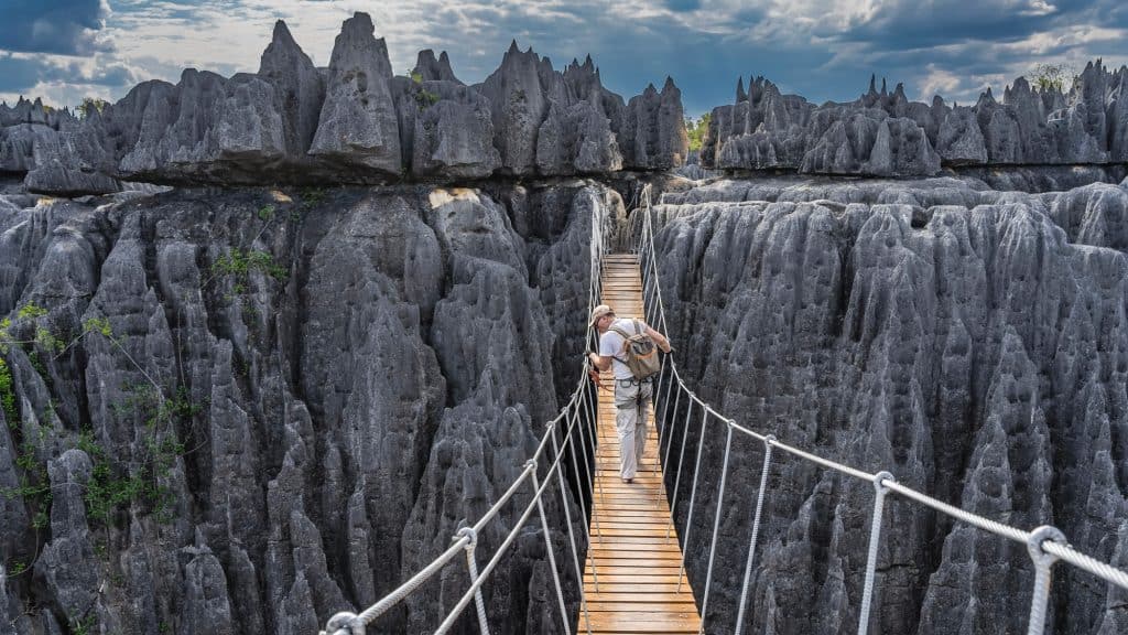 A hiker crosses a suspension bridge over sharp, jagged limestone formations under a partly cloudy sky, showcasing a dramatic and rugged landscape.